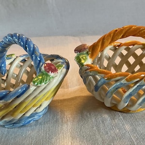 May include: Two ceramic decorative baskets with woven lattice designs. One basket has a blue handle and accents, the other has an orange handle and accents. Both have floral and leaf details.