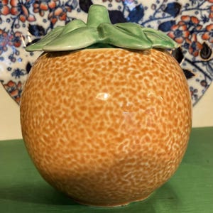 May include: A ceramic orange-shaped jar with a green leaf-shaped lid. The jar is a mottled orange colour, and the lid is a light green. The jar is on a green surface, with a decorative plate in the background.