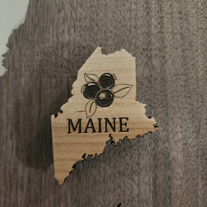 May include: A wooden cutout of the state of Maine with a design of blueberries and the word "MAINE" in black lettering.