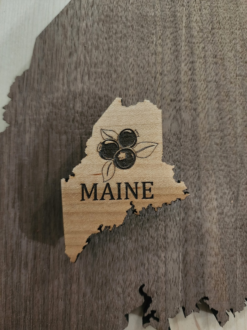 May include: A wooden cutout of the state of Maine with a design of blueberries and the word "MAINE" in black lettering.