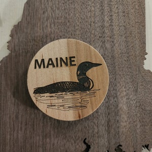 May include: A wooden round ornament with a black silhouette of a loon swimming on a lake. The word "MAINE" is engraved above the loon.