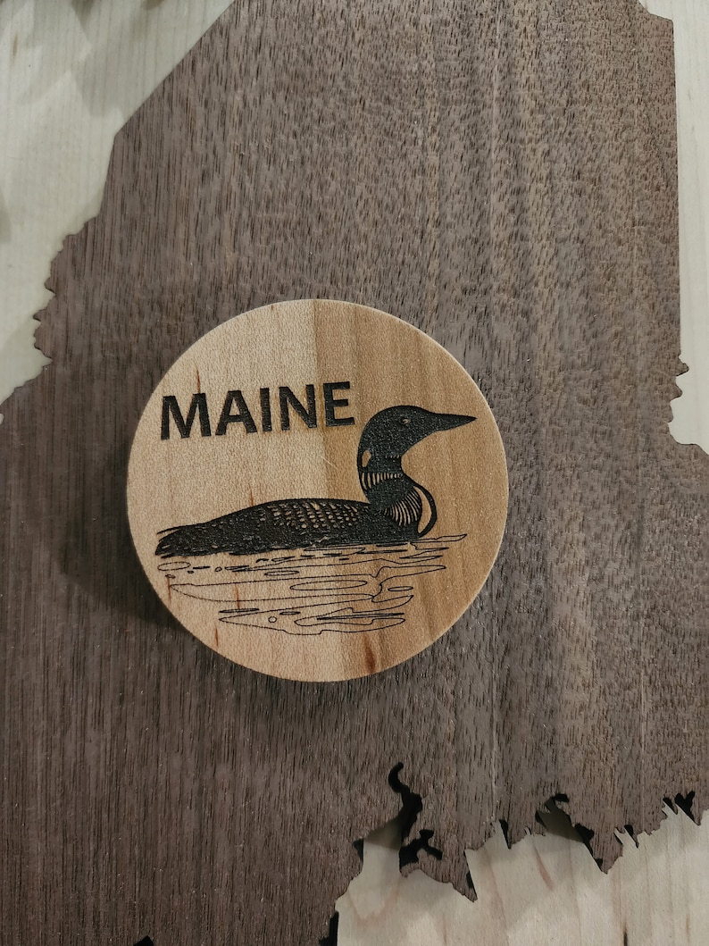 May include: A wooden round ornament with a black silhouette of a loon swimming on a lake. The word "MAINE" is engraved above the loon.