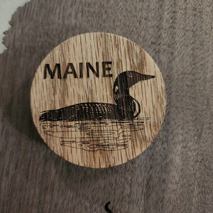 May include: A wooden coaster with a black and white image of a loon and the word "MAINE" burned into the wood.