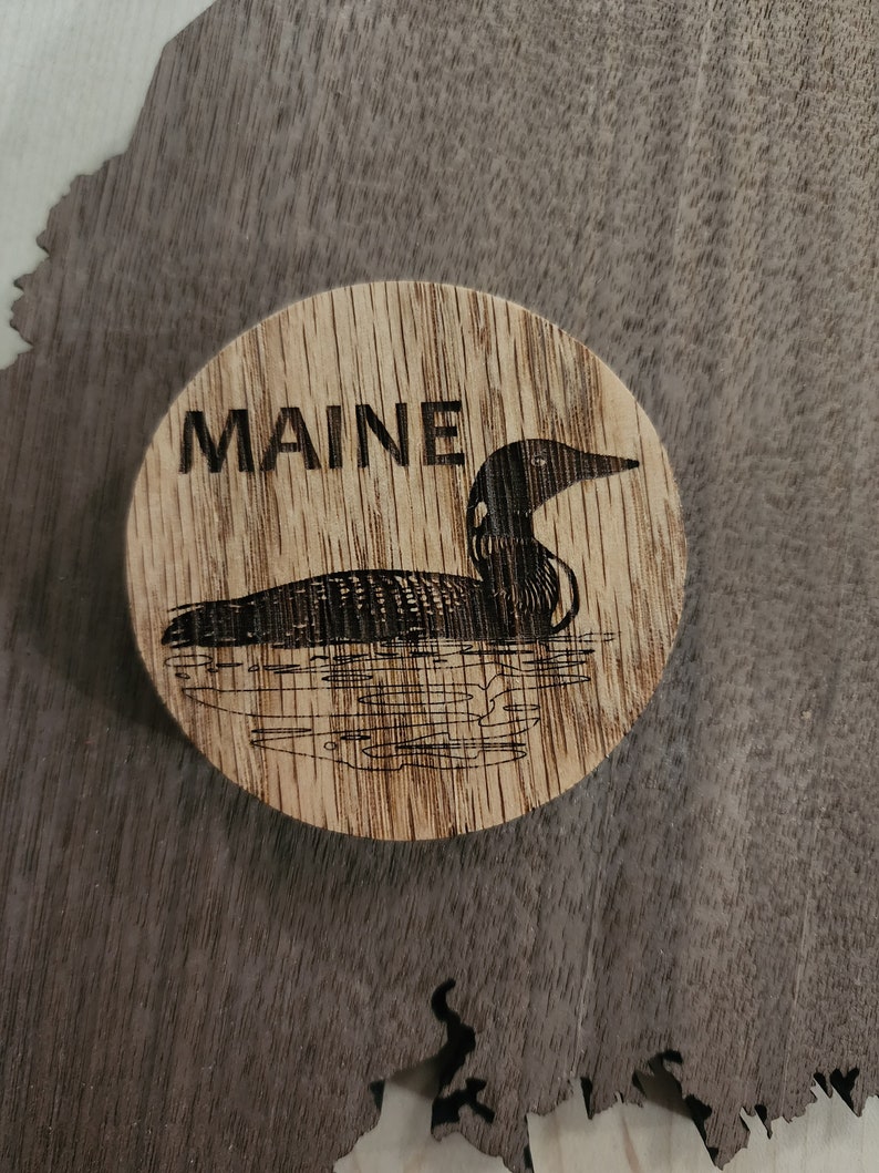 May include: A wooden coaster with a black and white image of a loon and the word "MAINE" burned into the wood.
