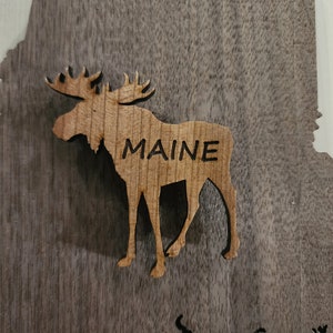 May include: A wooden cutout of a moose with the word "MAINE" carved into its side. The moose is standing on a dark brown wooden background.