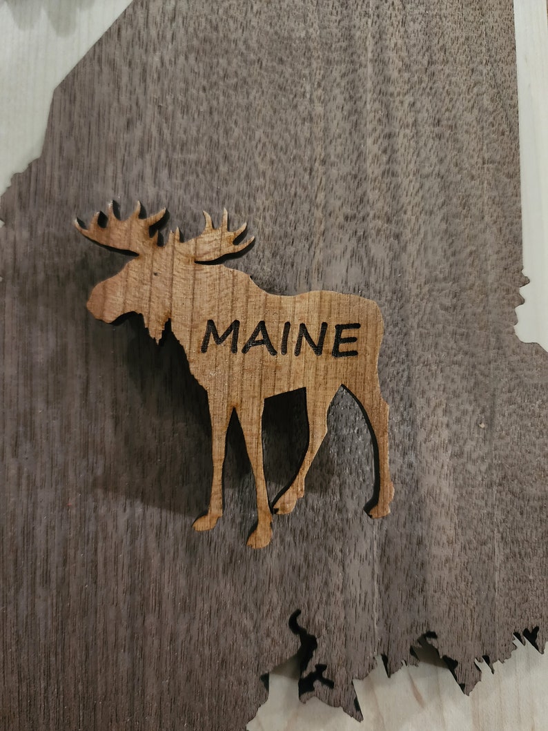 May include: A wooden cutout of a moose with the word "MAINE" carved into its side. The moose is standing on a dark brown wooden background.