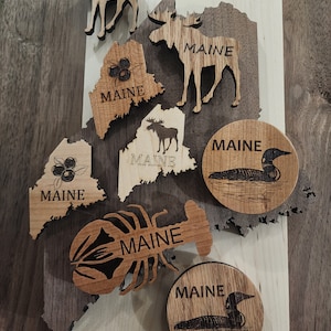 May include: A collection of wooden cutouts featuring the state of Maine, a moose, a lobster, a loon, and blueberries. Each cutout has the word "Maine" inscribed on it.