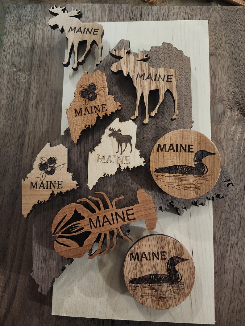 May include: A collection of wooden cutouts featuring the state of Maine, a moose, a lobster, a loon, and blueberries. Each cutout has the word "Maine" inscribed on it.