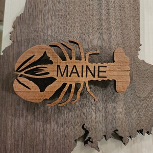 May include: A wooden cutout of a lobster with the word "MAINE" carved into it. The lobster is brown and the background is a dark wood grain.