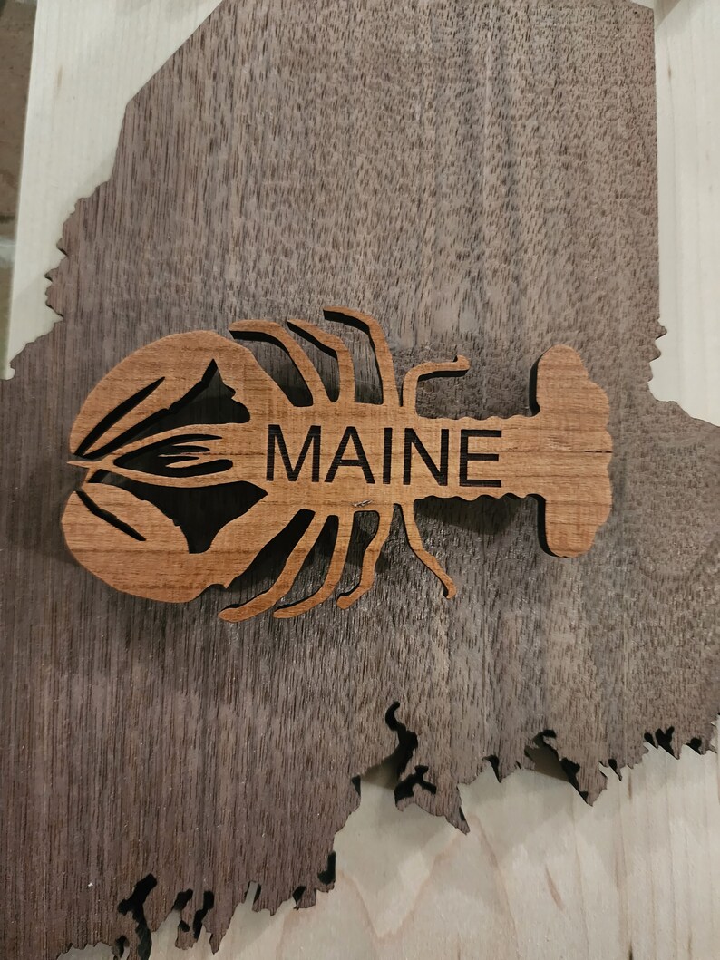 May include: A wooden cutout of a lobster with the word "MAINE" carved into it. The lobster is brown and the background is a dark wood grain.