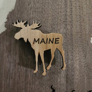 May include: A wooden cutout of a moose with the word "MAINE" carved into its side. The moose is standing on a dark brown wooden surface.