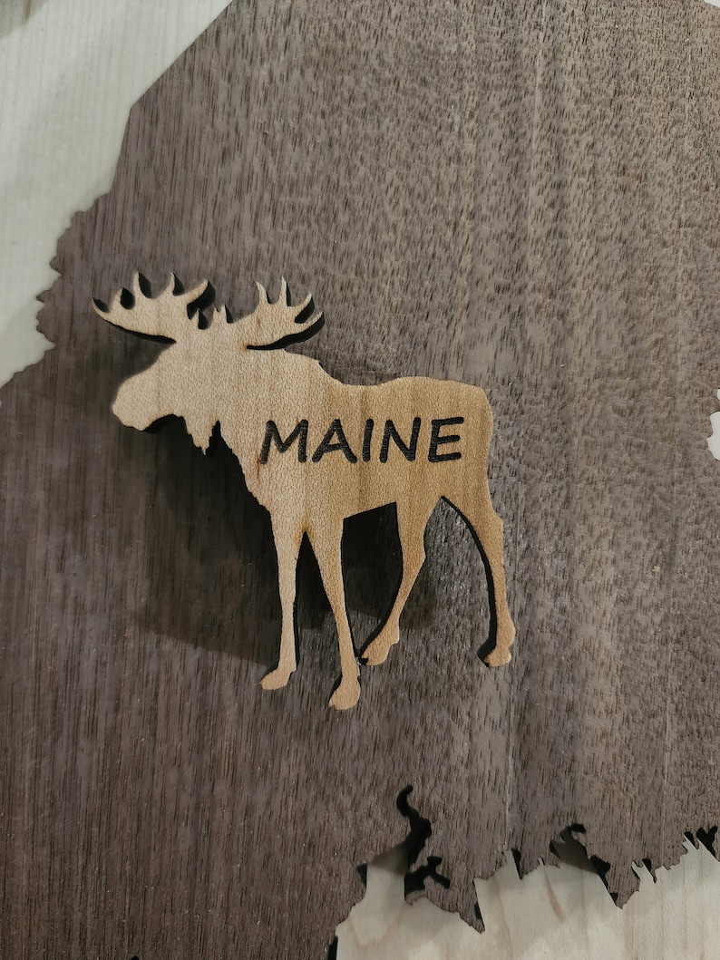 May include: A wooden cutout of a moose with the word "MAINE" carved into its side. The moose is standing on a dark brown wooden surface.