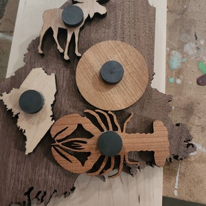 May include: A wooden cutout of the state of Maine with a moose, lobster, and a circle with a magnet in each cutout. The wood is a dark brown color.