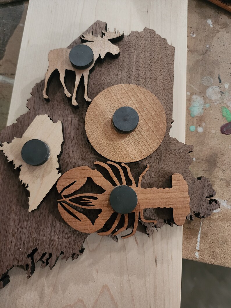 May include: A wooden cutout of the state of Maine with a moose, lobster, and a circle with a magnet in each cutout. The wood is a dark brown color.