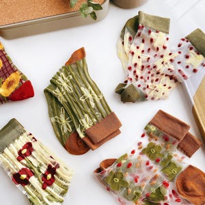 以下が含まれることがあります： 花柄の刺繍が施された透き通るような靴下5足セット。靴下は緑、茶色、赤の色合いで、繊細で女性らしい印象です。