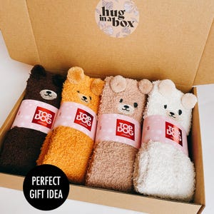 May include: Four pairs of fuzzy animal-themed socks in a cardboard gift box. Each pair features a bear face with ears and a pink paper band labelled Top Dog London. The box lid has a circular sticker that reads Hug in a Box.