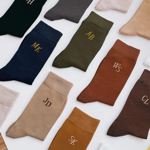 Op de afbeelding: Een collectie effen sokken in de kleuren zwart, marineblauw, olijfgroen, roest, bruin en beige. Elke sok is voorzien van geborduurde goudkleurige initialen, waaronder MK, AB, WS, JD, CL en SK, gerangschikt in een platte compositie.