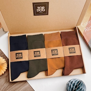 Puede incluir: Un conjunto de cuatro pares de calcetines en azul marino, verde oliva, amarillo mostaza y marrón, presentados en una caja de cartón. Cada par está sujeto con una banda de papel color canela con el texto negro Top Dog London impreso.