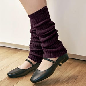 Puede incluir: Polainas de color púrpura oscuro con textura de punto acanalado. Se combinan con zapatos negros estilo Mary Jane, con una correa y hebilla. Los zapatos están sobre un suelo de madera.