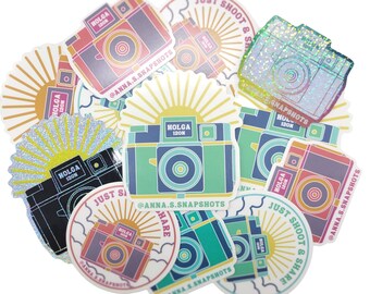 Pode incluir: Uma coleção de adesivos coloridos com um design de câmera vintage com o texto "Holga 120N" e a frase "Just Shoot & Share". Os adesivos têm um design de raios de sol e são perfeitos para adicionar um toque retrô aos seus pertences.