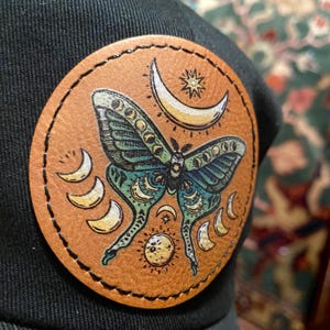 May include: A brown leather patch with a green and blue moth with moon and star designs. The patch is stitched around the edges.