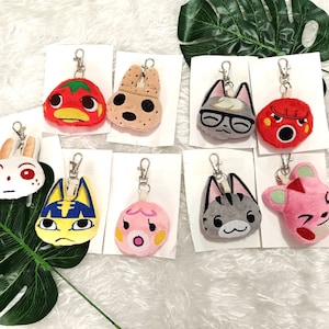 May include: A set of ten plush animal keychains featuring various characters. The keychains are attached to white cards with silver rings. The characters include a red tomato, a brown dog, a gray cat with glasses, a red and white fox, a white rabbit, a blue and yellow cat, a pink pig, a gray cat, a pink mouse, and a red and white fox.