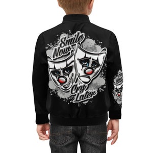 May include: Black bomber jacket with a white and gray graphic of two theatrical masks. The masks are labeled "Smile Now" and "Cry Later".