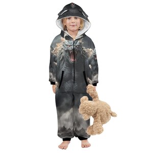 Op de afbeelding: Een kind draagt een grijze onesie met capuchon met een monsterontwerp. De onesie heeft een gedetailleerde illustratie van een brullend monster met rode ogen en een wit gevoerde capuchon met oren. Het kind houdt een bruine teddybeer vast.