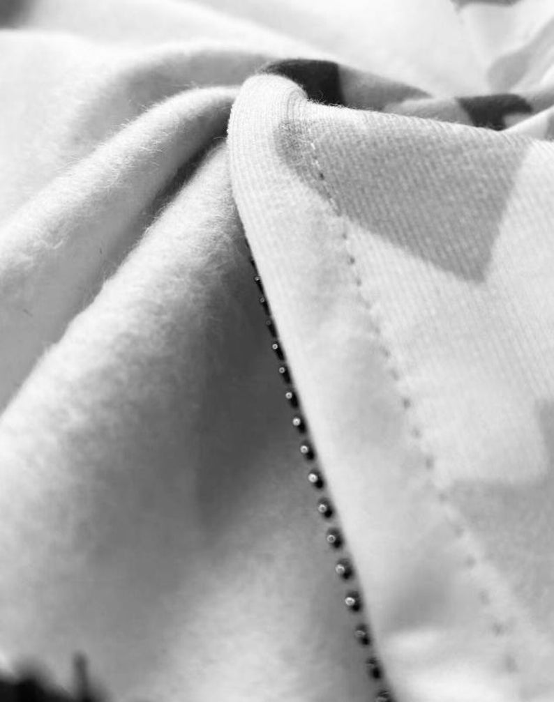 Op de afbeelding: Close-up van een witte stof met een ritsdetail. De stof is over zichzelf gevouwen, waardoor de stiksels en de rits tanden zichtbaar zijn.