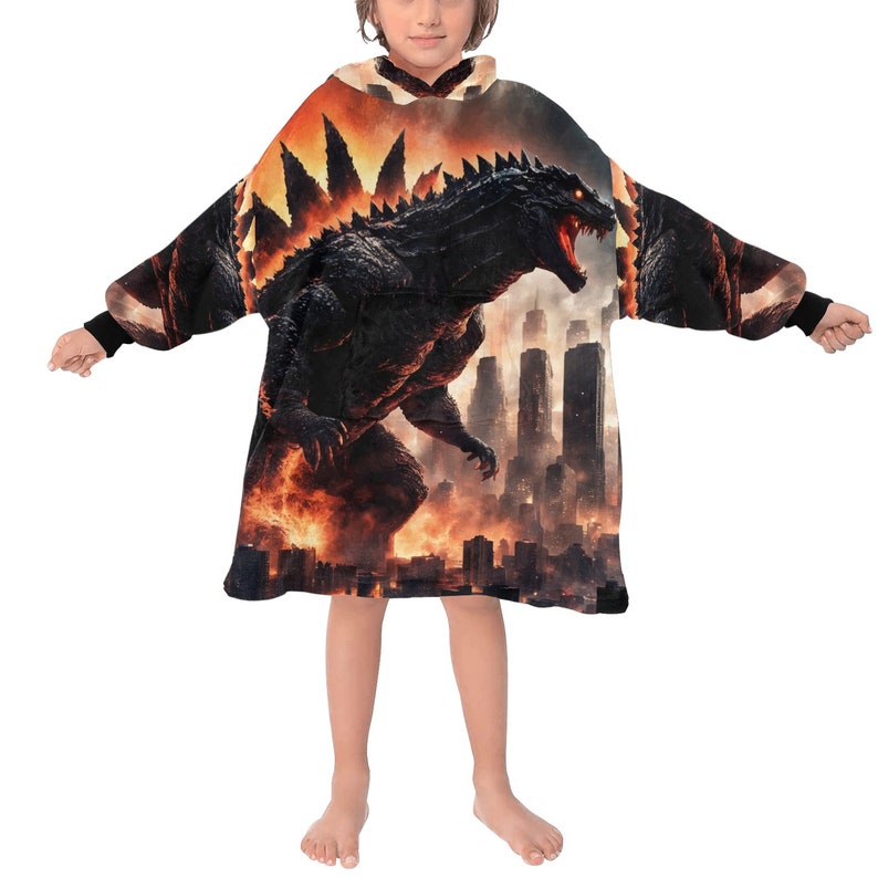 May include: A black and orange hooded blanket featuring a Godzilla graphic. The design shows Godzilla in a city setting with flames and skyscrapers. The blanket has long sleeves with black cuffs.