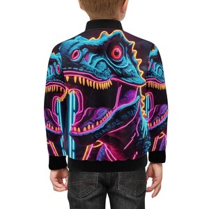 Könnte beinhalten: Eine schwarze Bomberjacke mit einem neonrosa, blauen und gelben Dinosaurier-Druck. Der Dinosaurier ist ein T-Rex mit offenem Maul und sichtbaren Zähnen. Die Jacke hat einen Reißverschluss und gerippte Bündchen und einen Bund.