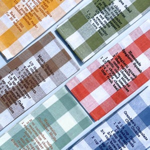 May include: Six kitchen towels with red, blue, green, and brown gingham patterns. Each towel features a handwritten recipe for a different dish, including Pea Salad, Homemade Spaghetti Squash, Grandma's Chocolate Chip Cookies, Mom's Chicken Noodle Soup, Watermelon Cucumber Mojito, and a recipe for eggs.