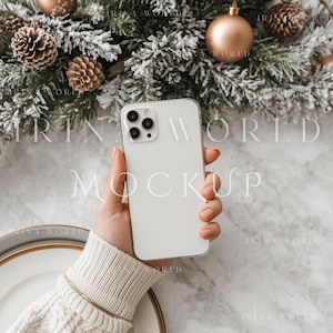 May include: A white smartphone held in a hand, with a blurred background of Christmas ornaments and frosted pine branches. The phone has a clear case and multiple camera lenses. A gold bracelet and a white sweater are also visible.