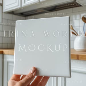 Puede incluir: Una maqueta de baldosa de cerámica blanca cuadrada sostenida por una mano. La baldosa tiene las palabras "IRINA WORLD MOCKUP" en una fuente clara y elegante. El fondo muestra un entorno de cocina con gabinetes blancos y encimeras de madera.