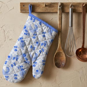 May include: A quilted oven mitt featuring a light blue and white chequered pattern with a repeating print of blue croissants on plates and blue leafy sprigs. The mitt has a solid blue fabric trim along the top edge and a matching blue hanging loop.