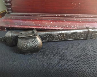 May include: A vintage, ornate, metal inkwell with a hinged lid and intricate floral designs. The inkwell is made of dark metal and has a worn, antique finish.