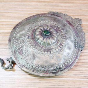 Puede incluir: Un objeto decorativo redondo de color plateado con diseños intrincados y una piedra preciosa verde en el centro. El objeto tiene un gancho en el lateral.