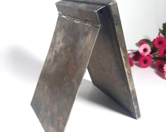 May include: A silver-toned, hinged, folding mirror with a worn, textured surface. The mirror is standing upright on a white surface.