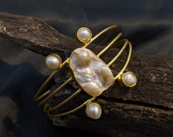 Puede incluir: Un brazalete dorado con una perla grande de forma irregular en el centro, rodeada de cuatro perlas más pequeñas. El brazalete tiene un diseño de múltiples hebras y se muestra sobre un trozo de madera oscura sobre un fondo oscuro.