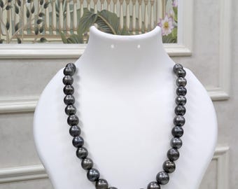 May include: A dark gray pearl necklace displayed on a white mannequin bust. The pearls are round and evenly sized, with a subtle luster. The necklace is a classic jewelry piece, suitable for formal occasions.