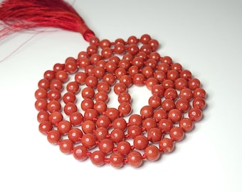 Puede incluir: Un collar de cuentas mala rojo con borla. El collar está hecho de cuentas redondas de color marrón rojizo ensartadas en un cordón rojo. La borla es de color rojo intenso.