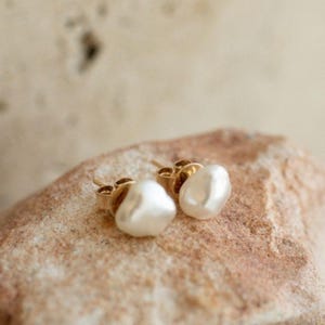 Op de afbeelding: Een paar gouden oorstekers met onregelmatige, witte pareldruppels. De oorbellen zijn geplaatst tegen een neutrale, getextureerde achtergrond, die de natuurlijke vorm en glans van de parels benadrukt. De gouden zetting voegt een vleugje elegantie toe.