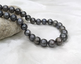 May include: A strand of dark gray pearls, each with a lustrous sheen, rests on a light-colored surface. The pearls are round and evenly sized, creating a classic jewelry piece. The necklace is draped over a textured, light-colored stone.