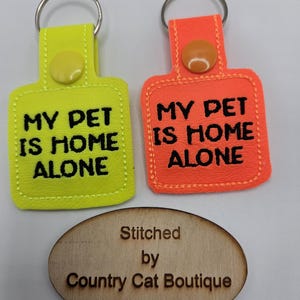 May include: Two keychains with the text "MY PET IS HOME ALONE" in black. One keychain is neon yellow with a yellow button, and the other is orange with an orange button. A wooden oval with the text "Stitched by Country Cat Boutique" is below.