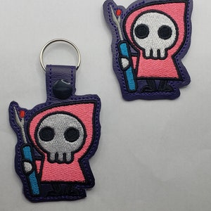 May include: Two embroidered keychains featuring a cartoon grim reaper design. The reaper is in a pink hooded robe with a skull face, holding a blue and teal tool. The keychains have purple backing and a silver clasp.