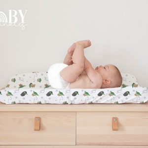 Op de afbeelding: Een witte verschoningsmatrashoes met een groene en bruine print van een cartoonfiguur. Een baby ligt met zijn benen in de lucht op de verschoningsmatras.