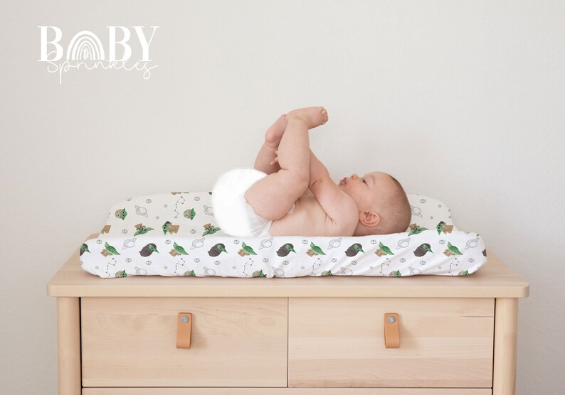Op de afbeelding: Een witte verschoningsmatrashoes met een groene en bruine print van een cartoonfiguur. Een baby ligt met zijn benen in de lucht op de verschoningsmatras.