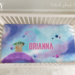 May include: A fitted crib sheet with a galaxy print featuring a green alien character holding a microphone. The sheet is blue, purple, and pink with white stars and planets. The name "BRIANNA" is printed in pink letters on the sheet.