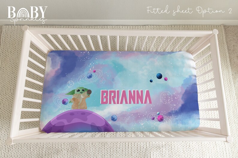 May include: A fitted crib sheet with a galaxy print featuring a green alien character holding a microphone. The sheet is blue, purple, and pink with white stars and planets. The name "BRIANNA" is printed in pink letters on the sheet.