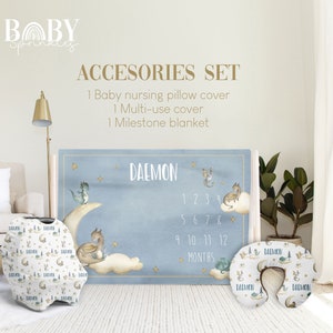 Puede incluir: Un conjunto de accesorios para bebé con una manta de hitos azul y blanca, una funda para cojín de lactancia y una funda multiusos. La manta presenta una luna, nubes y dragones con el nombre "DAEMON" y los números del 1 al 12 para los meses. Las fundas presentan un patrón repetido de dragones, lunas y estrellas.